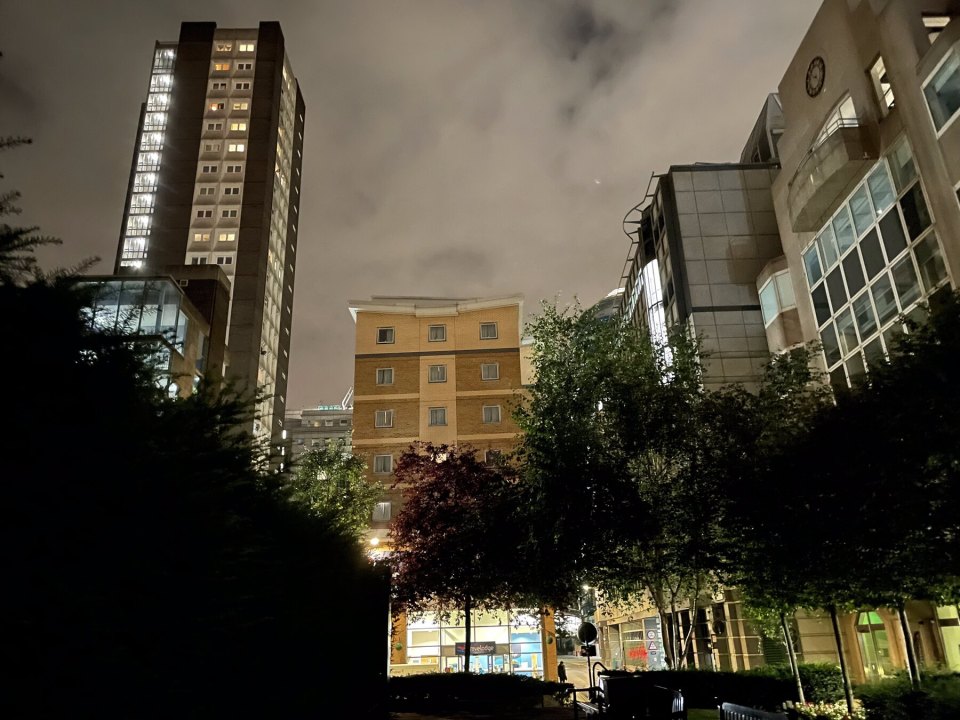 A lit residential tower block in the City of London in the evening, trees in the courtyard