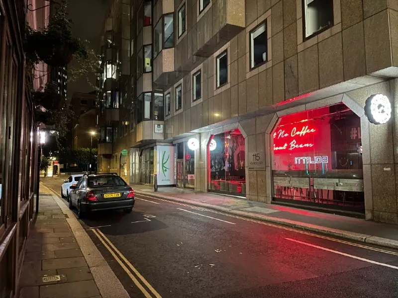 A dark saloon car parked at the kerb on a London street at night with a red neon café sign