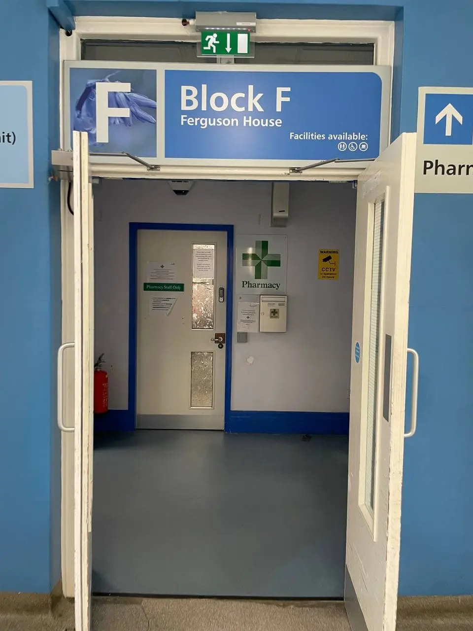 Коридор больницы NHS: синий указатель Block F Ferguson House над открытой дверью и вывеска Pharmacy с зелёным крестом