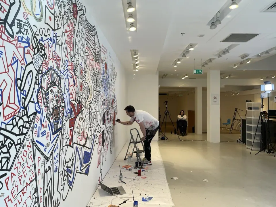 An artist on a stepladder painting a large white wall, paints and a laptop on the floor, cameras set up nearby